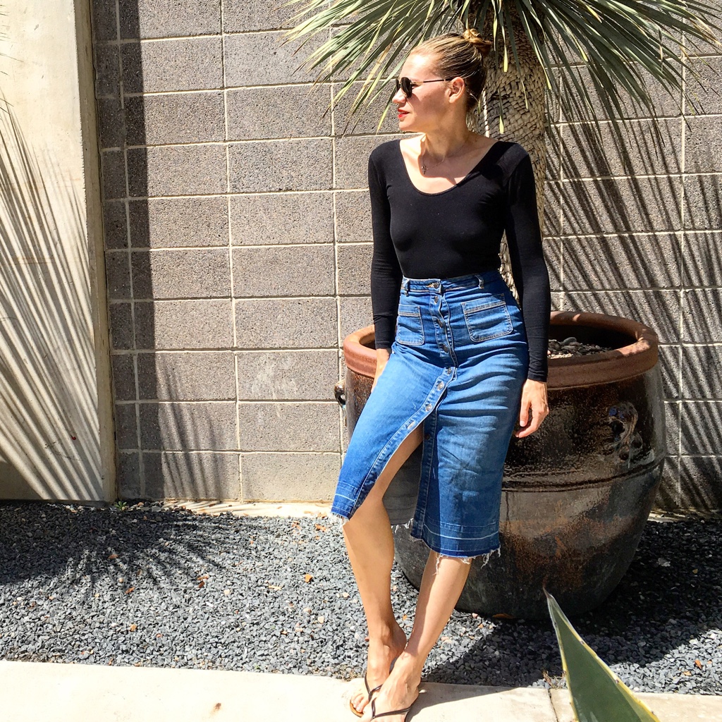 A woman in a black long sleeve leotard and a button up midi denim skirt leans against a huge planter