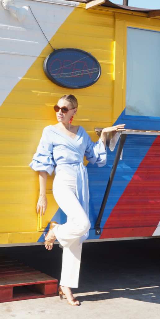 A woman in white pants, a blue and white wrap top in front of a red, white, blue, and yellow food truck 