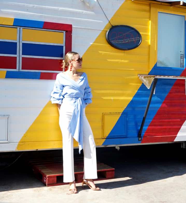 a woman in white pants and a wrap blue and white top stands in front of a white, red, blue, yellow food truck
