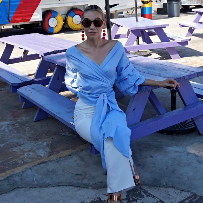 a woman in white pants and a blue and white wrap top sits at a picnic table in front of a red, white, blue, yellow food truck 