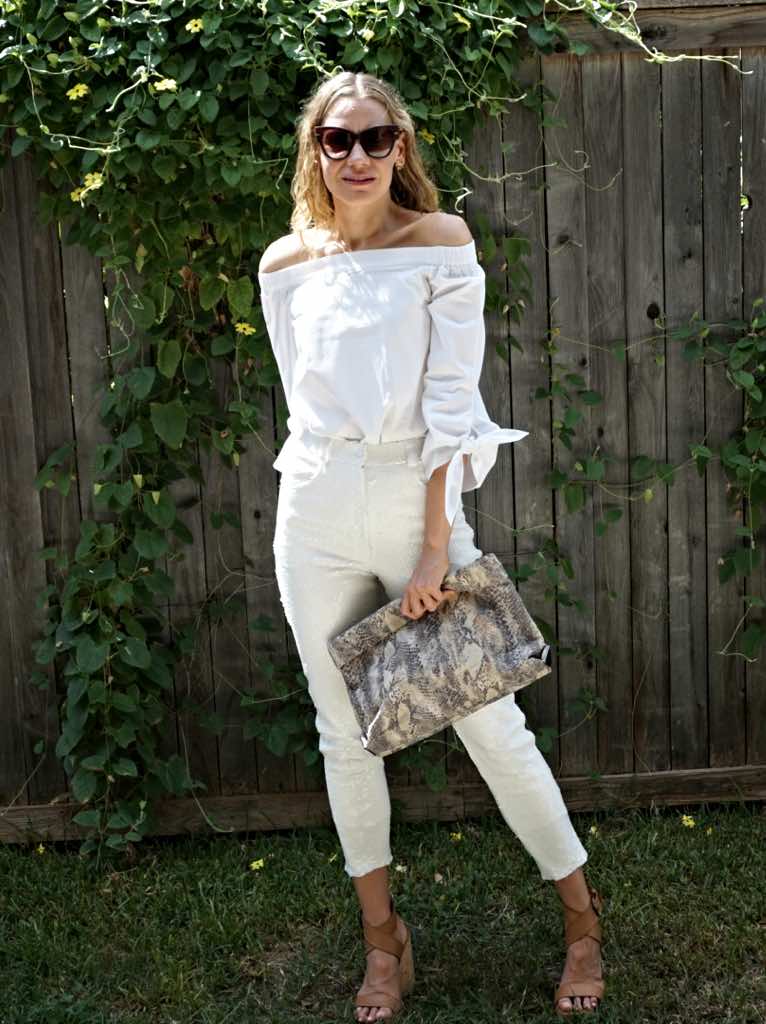 a woman in white pants and a white top with wedge sandals and a snake clutch in front of a fence