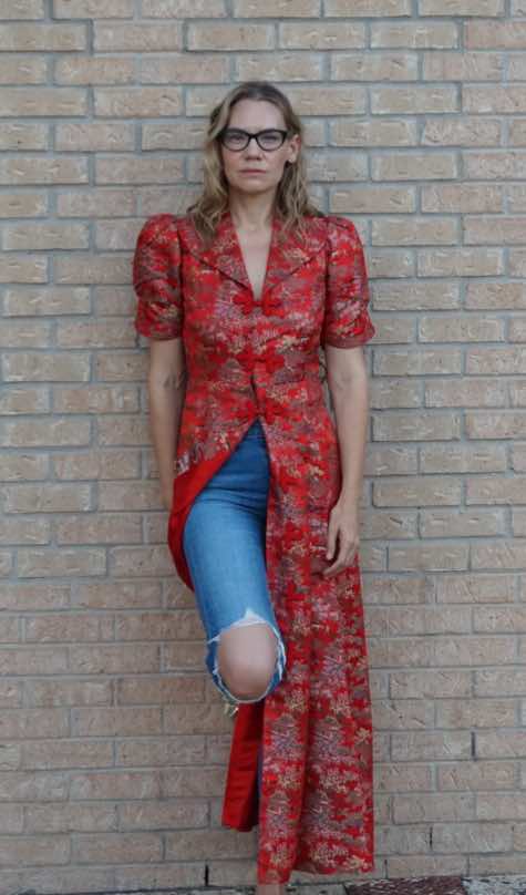 a woman in a red and gold print dress worn over jeans against a wall