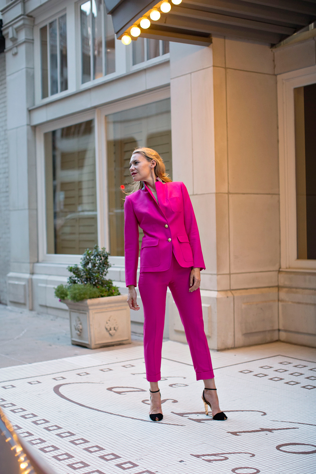a woman in a hot pink suit blazer and pants with black and gold heels in front a hotel door 