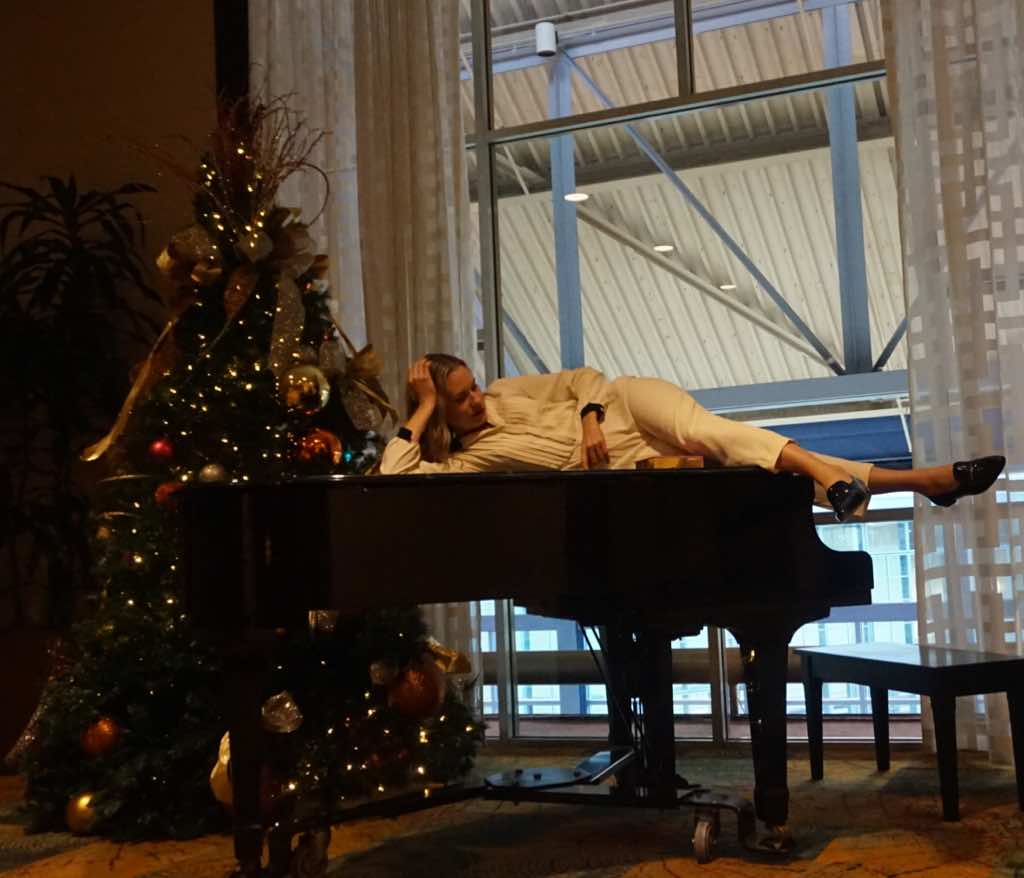 A woman in a white jumpsuit lounges on a piano by a Christmas tree