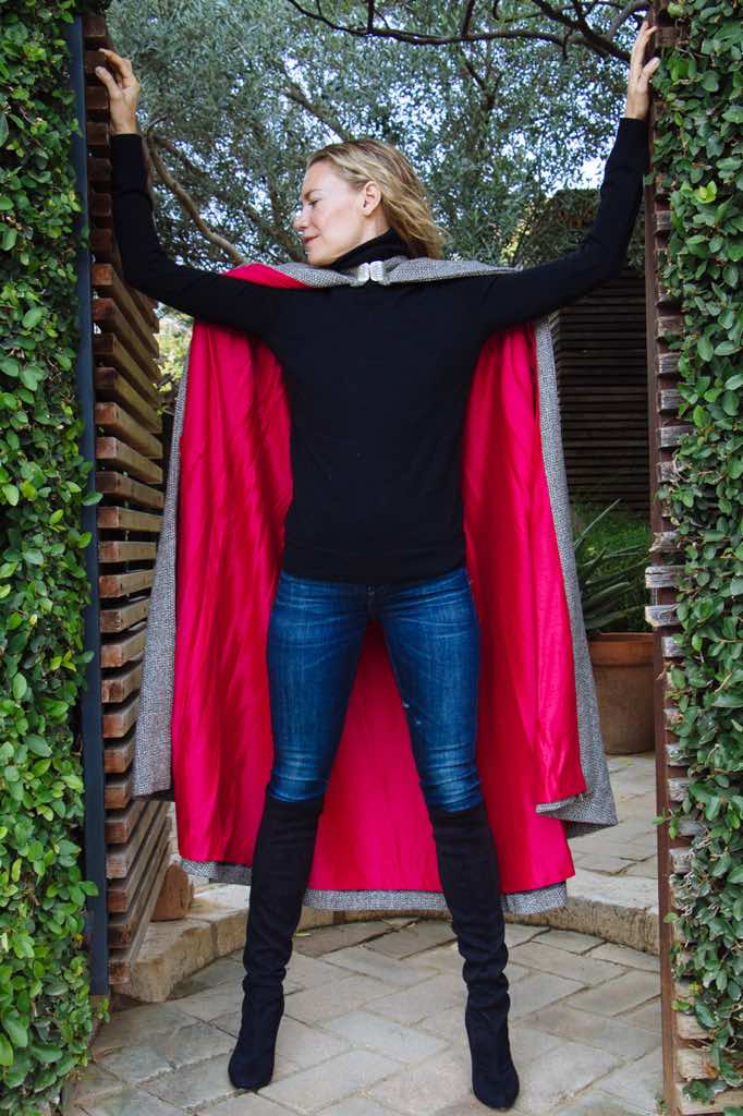 a woman in a garden doorway wearing a black turtleneck, jeans, boots and a pink lined maxi-cape