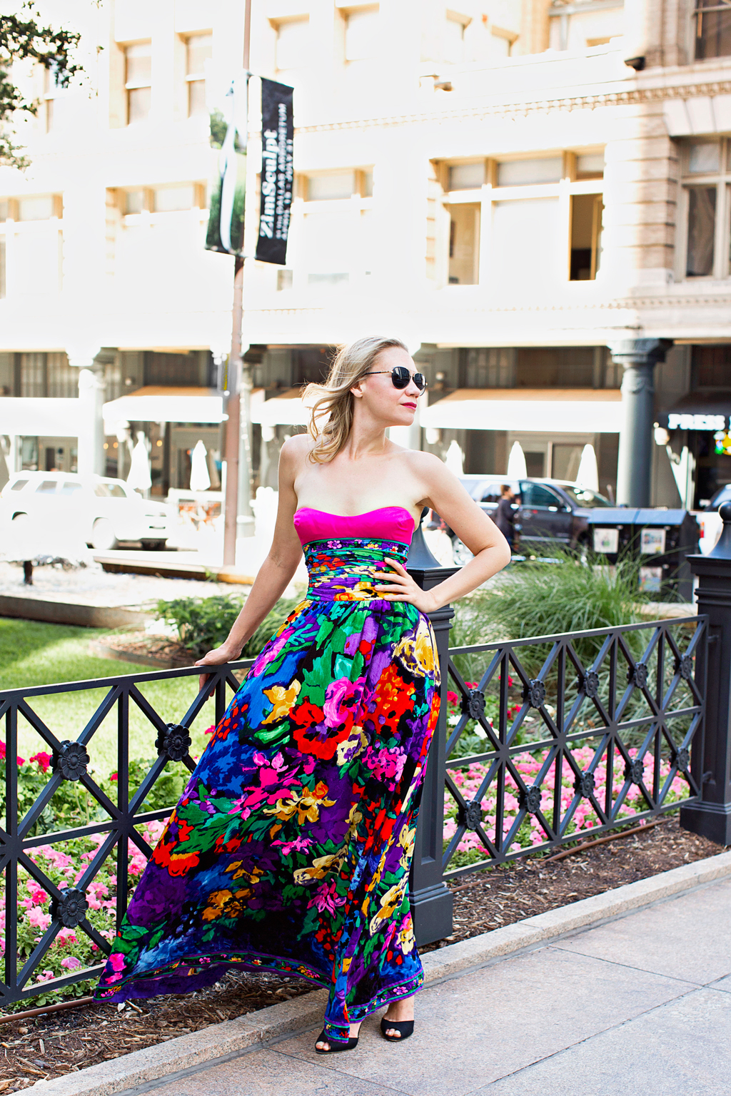 A blonde woman in a pink, blue, green and blue velvet gown in front of a wrought iron gate and green space 