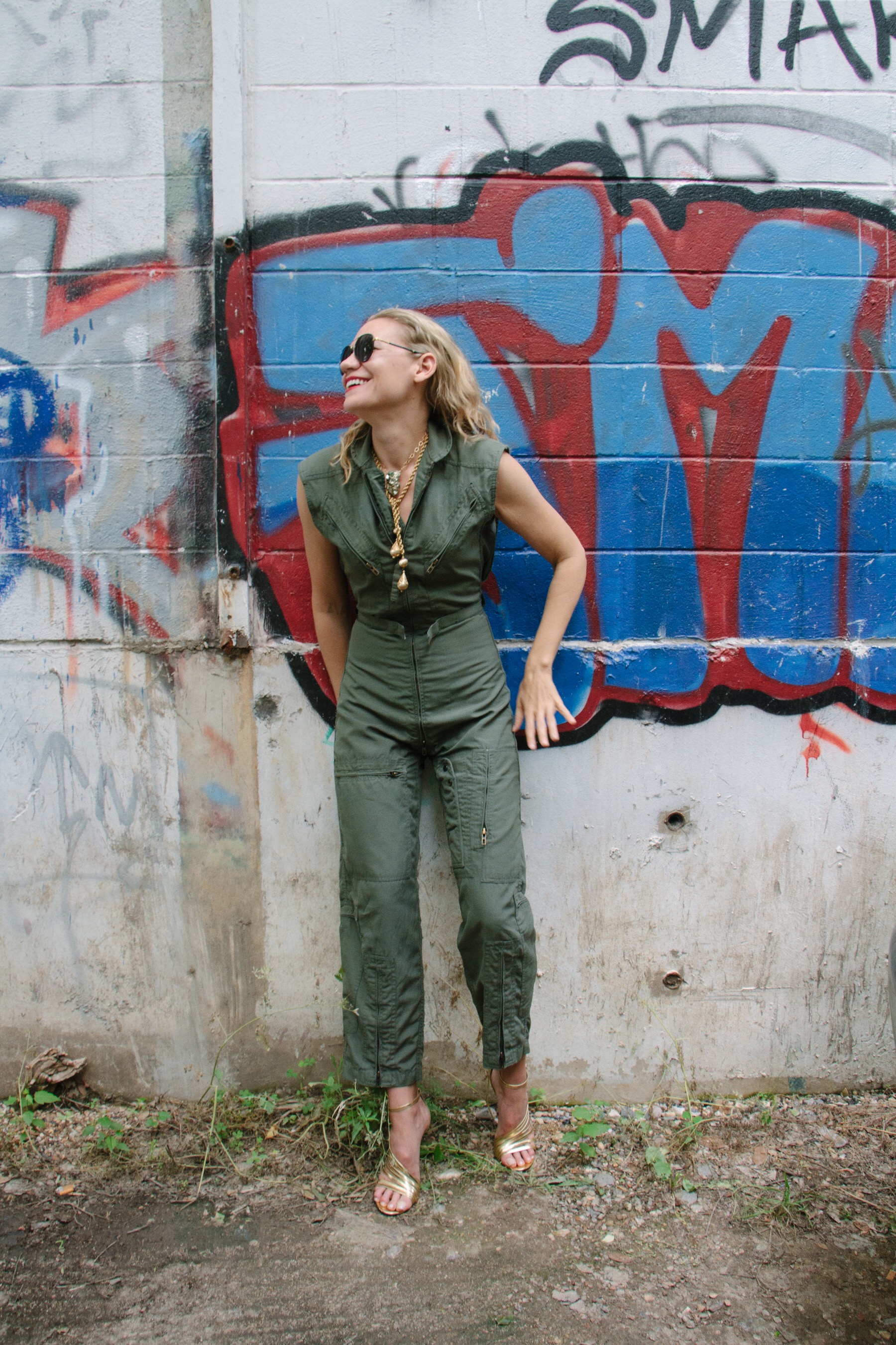 a blonde woman in a green flight suit, gold heels and necklaces in front of a concrete wall with graffiti 