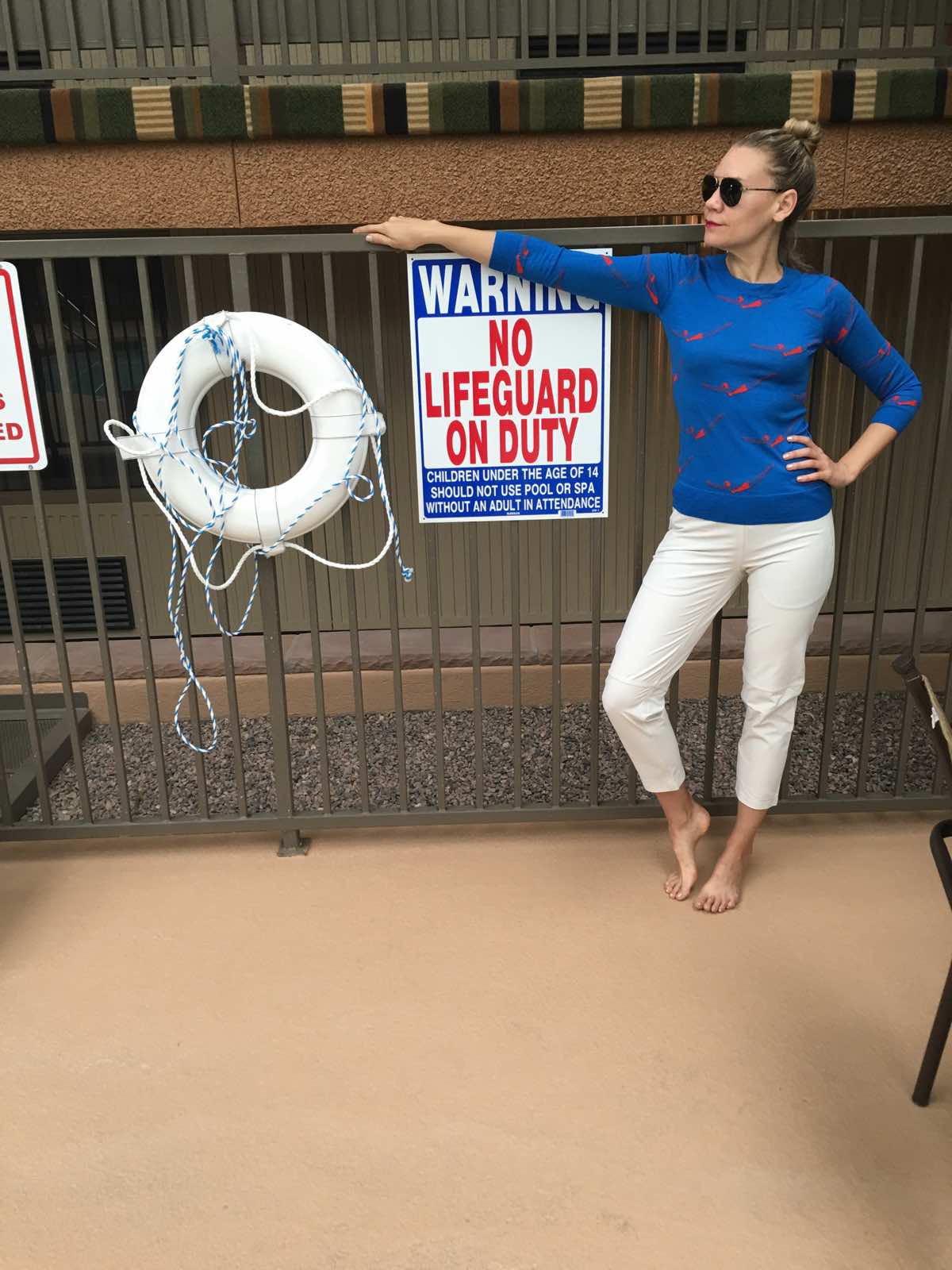 A woman in white pants and a blue sweater stands in front of a lifeguard sign