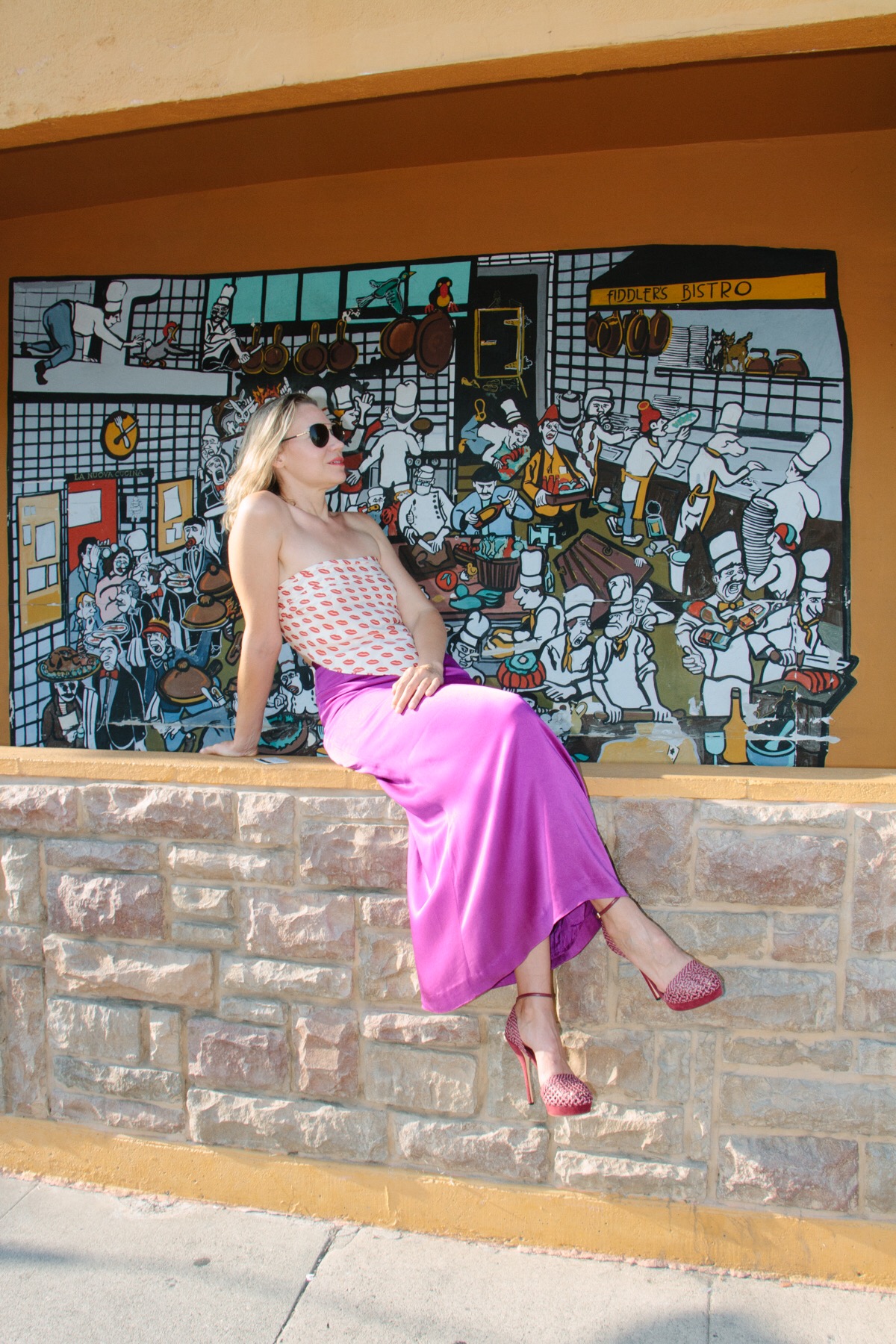 A woman in a kiss printed scarf and a purple maxi skirt sits on a stone wall
