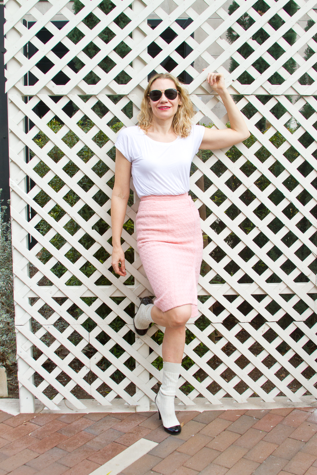 a woman in a white teeshirt, pink Chanel Boulce skirt, and white and black boots leans against a white trelice 