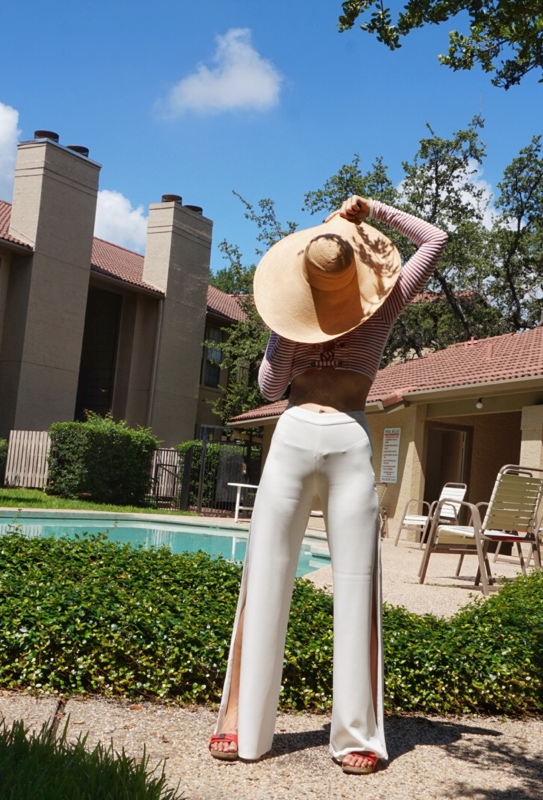 A woman in white pants, a red and white striped shirt holds a big hat over her face as she stands in front of a pool