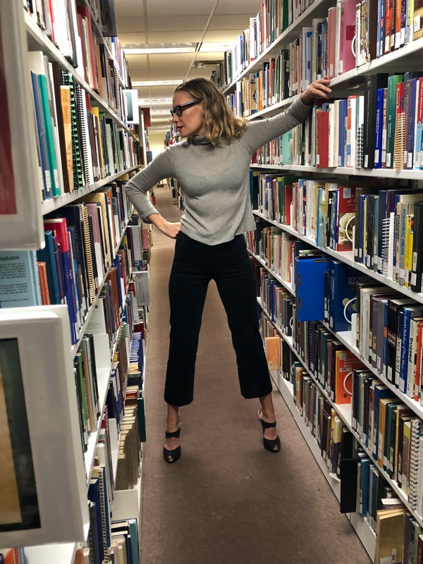 a woman in a grey sweater and dark corduroy pants stands in a row of bookshelves 