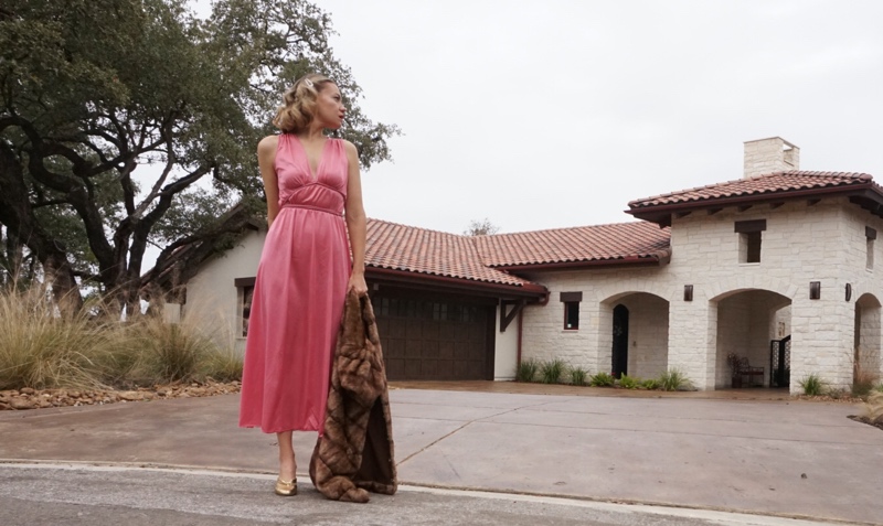 a woman in a pink dress holding a fur jacket in front of a house 