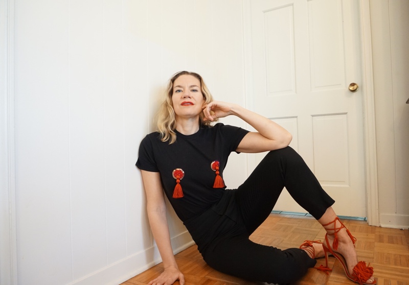 a woman in black tee shirt with tassels on it, black pants and red heels sits against a white wall