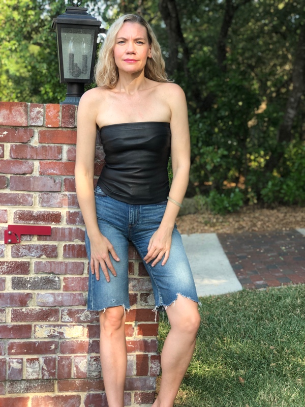 A blonde woman in jean shorts and a black leather tube top leaning against a stone wall