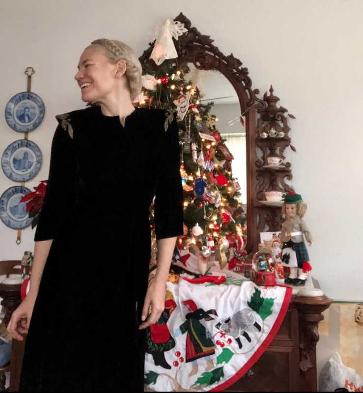 a blonde woman in a black velvet dress in front of a dresser with a small Christmas tree on top of it 