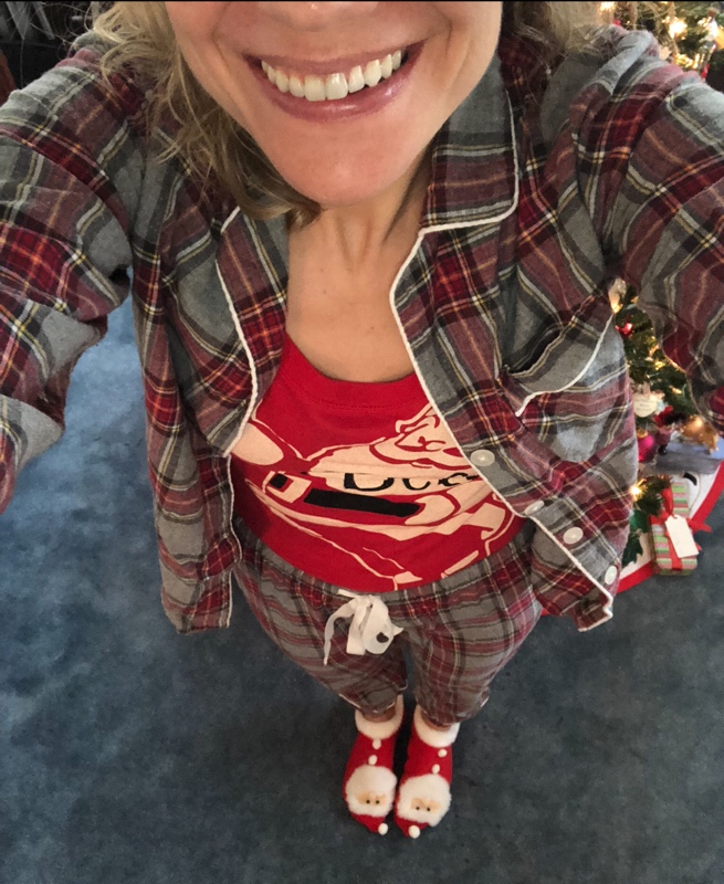A woman in grey plaid pjs and a Santa shirt with Santa slippers on