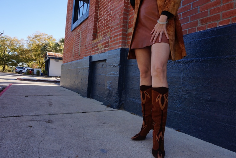 a woman in a brown leather mini skirt, a brown suede jacket and knee high boots in front of a brick wall