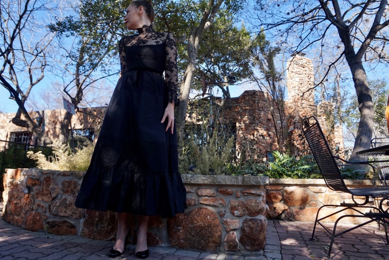 a woman in a black lace dress in front of a stone wall and trees
