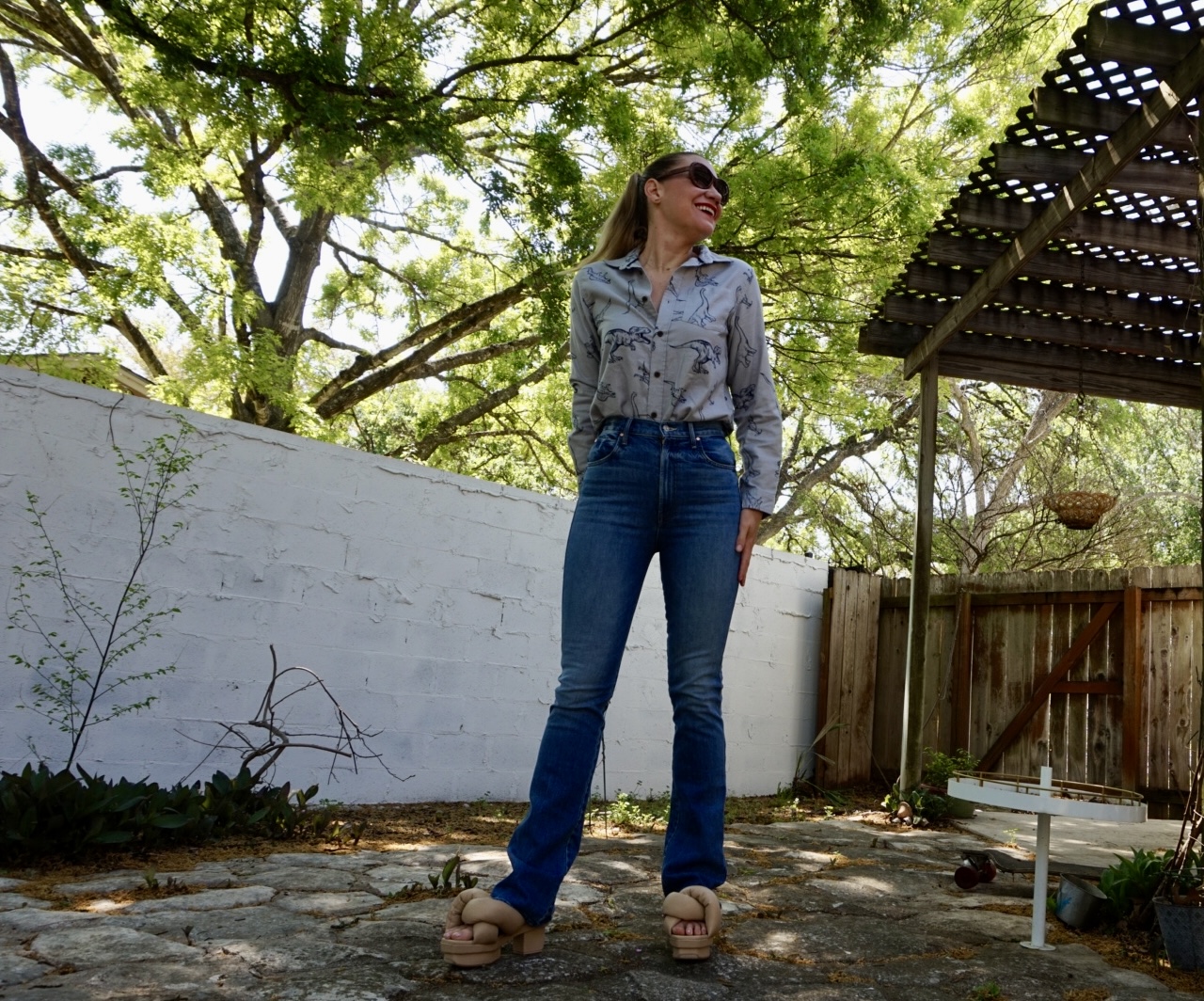a woman in a Dino printed button down and jeans with cream platform sandals on in front of a white fence