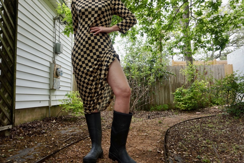 a woman in a checked dress and rain boots checking for rain in a green garden surrounded by a fence and a white wall