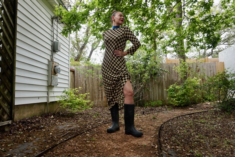 a woman in a checked dress and rain boots checks for rain in a green garden
