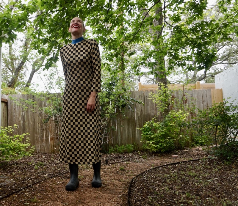 a woman in a checked dress and rain boots checks for rain in a green garden