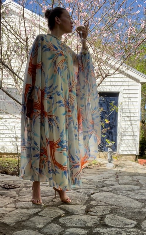a woman in a sheer caftan with orange and blue flowers on it stands in front of a blooming tree and a white house