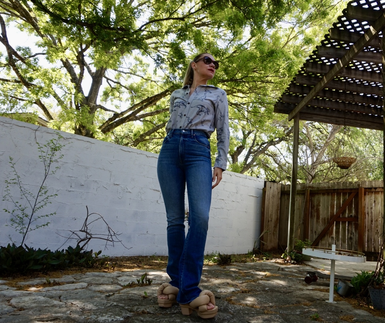a woman in a Dino print shirt and jeans with sandals in front of a white fence