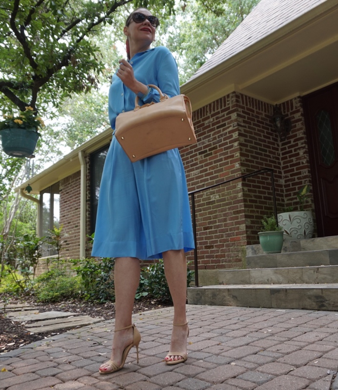 woman in a blue shirt and coulette set with a scarf in her hair and a satchel bag in front of a brick house