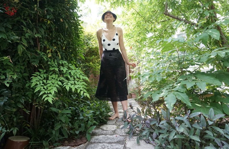 a woman in a lush green setting wears a white with black polka dot bathing suit with a sheer black skirt over and a straw hat