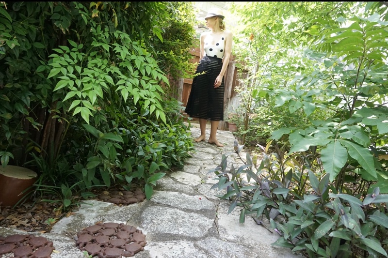 a woman in a lush green setting wears a white with black polka dot bathing suit with a sheer black skirt over and a straw hat