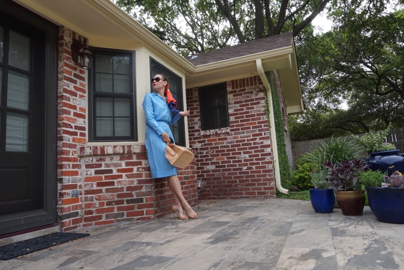 a woman in a blue silk shirt and short set with a scarf and a satchel bag in front of a brick house