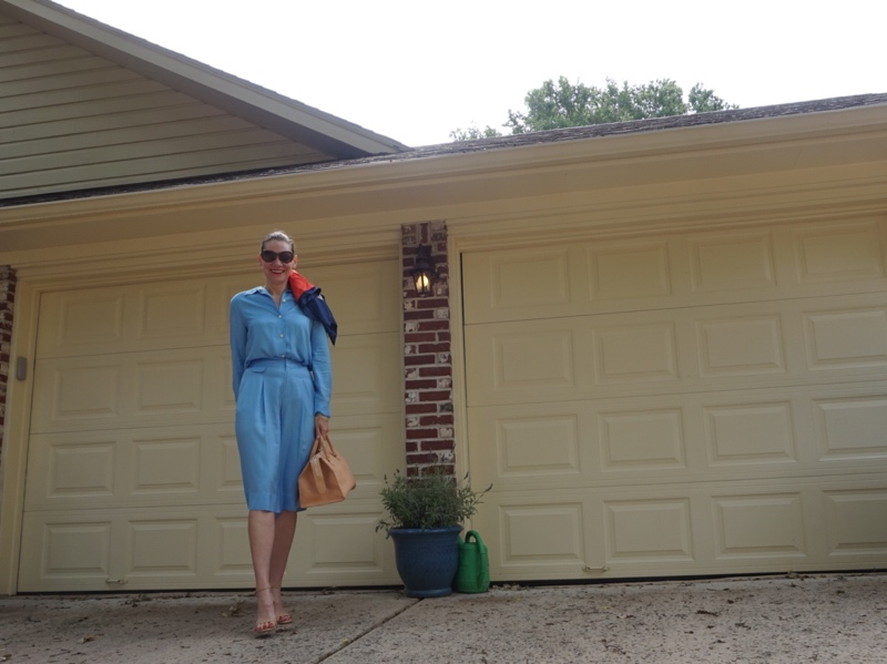 a woman in a blue silk short set with a scarf in her hair and her satchel bag walks out of a cream garage and brick house