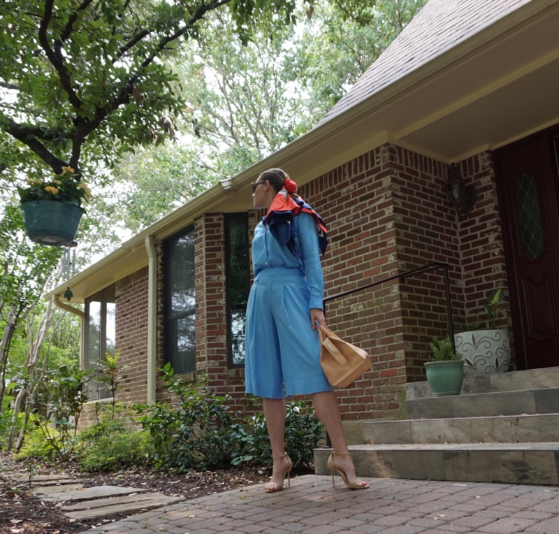 a woman in a blue silk set with a scarf in her hair and a satchel bag in front of a brick house