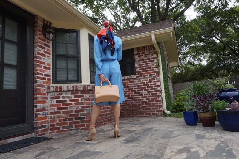 the black of a woman with a scarf in her hair and a satchel bag in front of a brick house