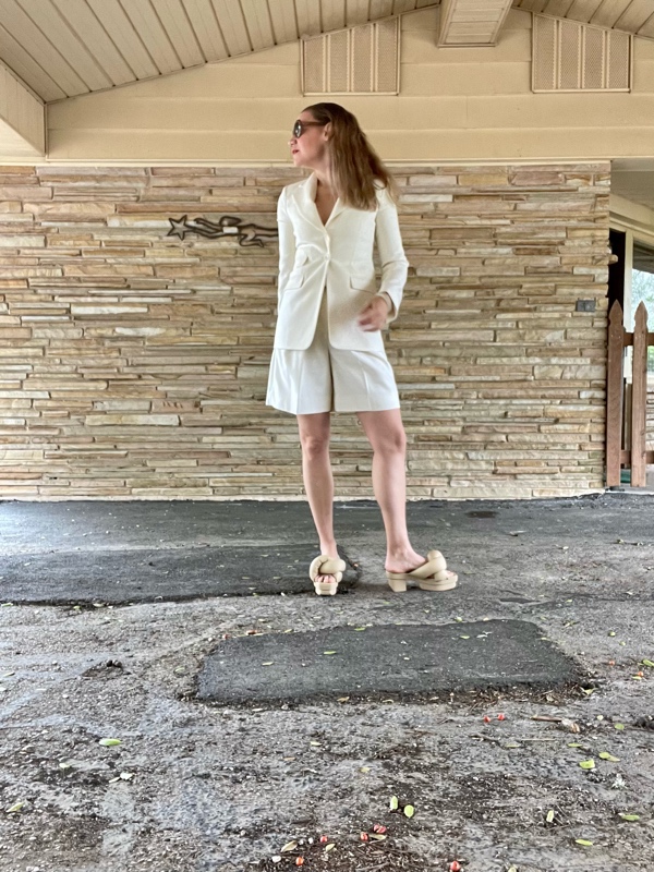 a woman in white shorts and a white blazer in front of a stone wall