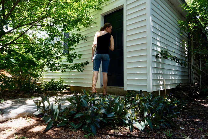 a woman in jean shorts and a blank one shoulder tank with lace up sandals stands in front of a white building, blue door and green trees