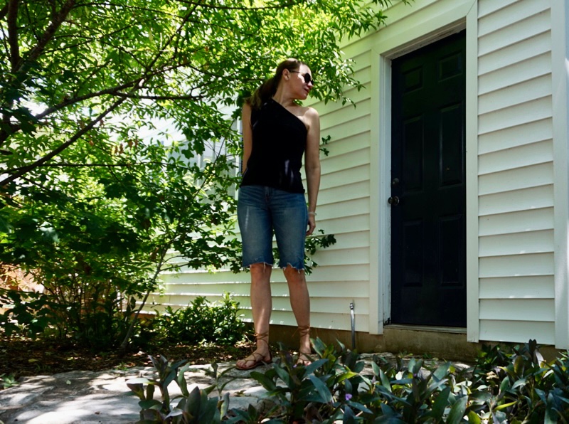 a woman in jean shorts and a blank one shoulder tank with lace up sandals stands in front of a white building, blue door and green trees