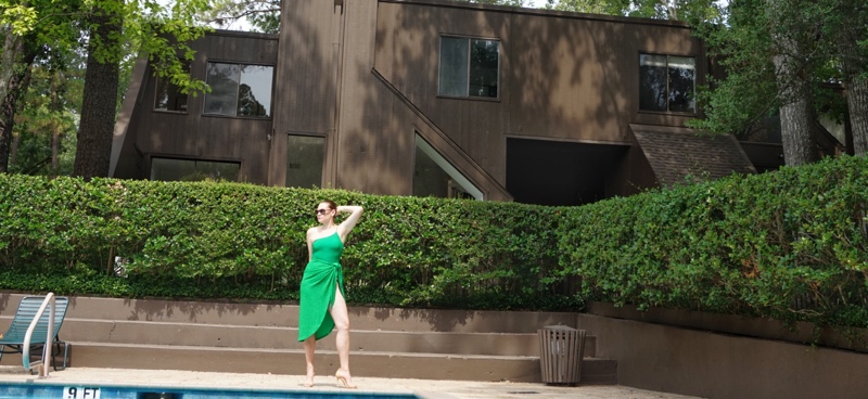 a woman in green one piece one shoulder sparkle swim and matching sarong with nude heels with a pool and green hedges and pool