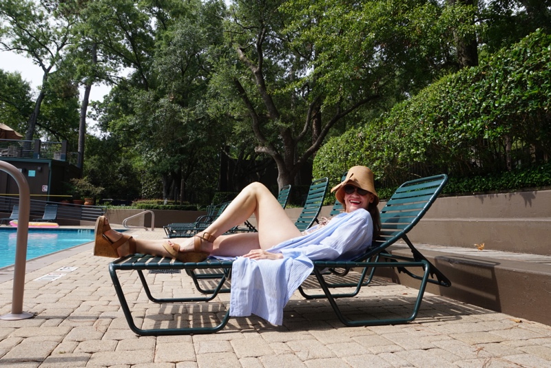 a woman lounges on a chair by the pool in a striped shirt dress, wedges, a bucket straw hat in front of a pool and green hedges 