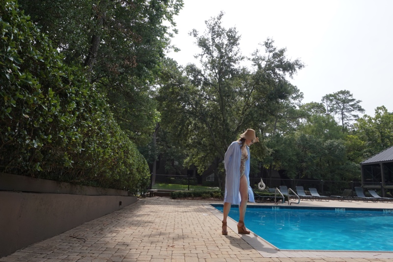 a woman in snakeskin one piece swimsuit and wedges blue and white shirt dress and bucket hat in front of a pool and hedge