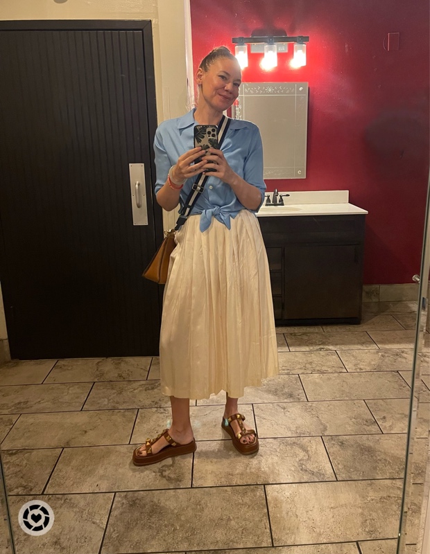 a woman in a blue silt shirt, white pleated silk skirt, studded brown sandals stands in front of a red wall, brown door, sink