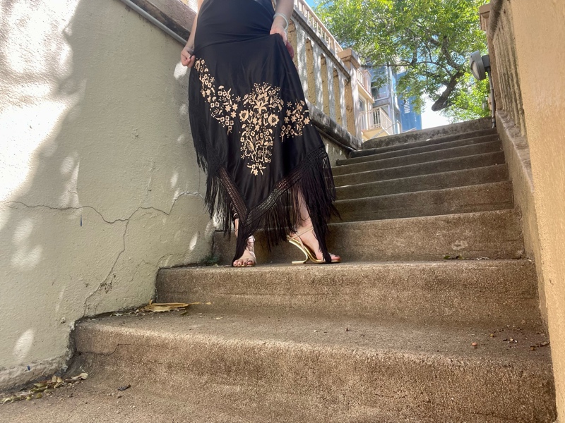 a woman in a strapless black dress and shawl with fringe and purse with fringe stands on stone steps