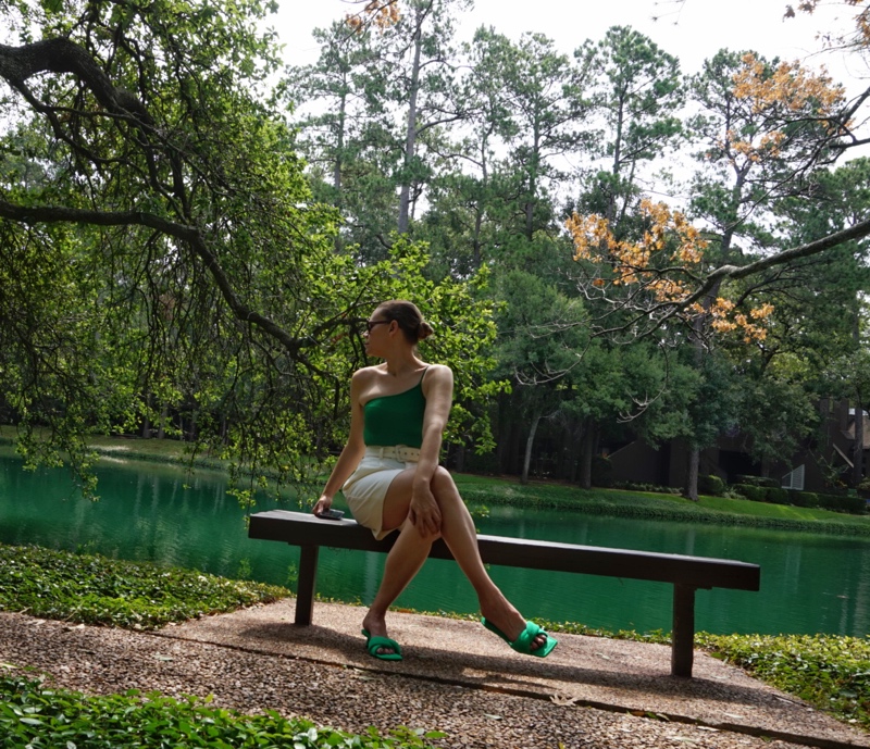 a woman sits on a bench in front of a lake in white shorts and a one shoulder green sparkle swim suit and green heels