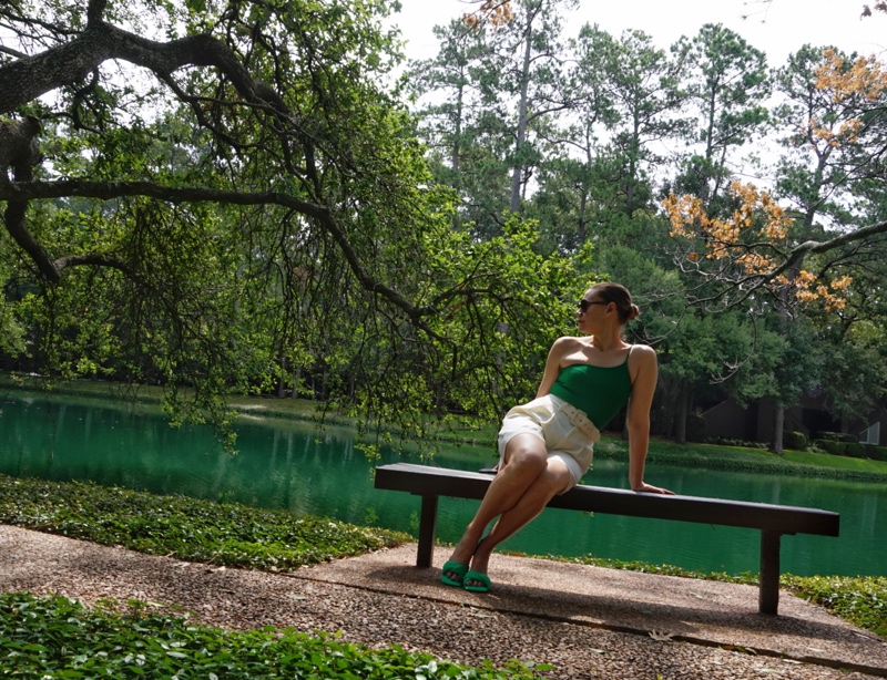 a woman lounging on a bench in front of a lake in a pair of white shorts, a green one shoulder body suit and green mules