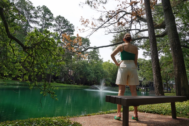 the back of a woman in white shorts and a green sparkle one piece swim suit in font of  a bench and a lake
