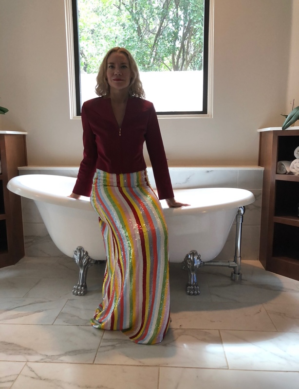 a blonde woman in a red jacket and a sequined striped maxi skirt sits on the edge of a white bathtub