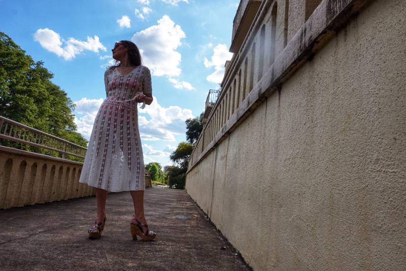 a woman in a white crochet dress with a bright swimsuit underneath walks on a stone walkway