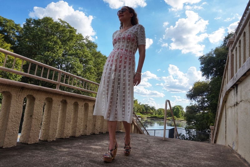 a woman in a white crochet dress with a bright swimsuit underneath walks on a stone walkway